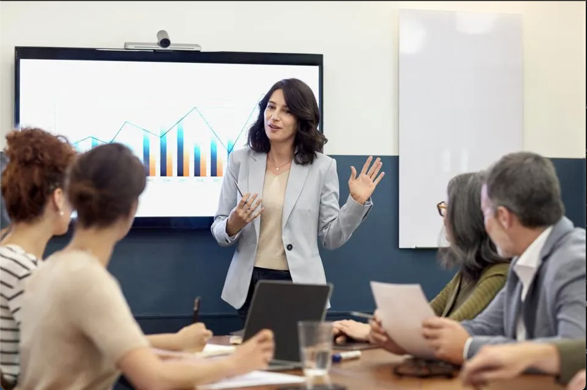 A woman in business clothing presenting graphs to a table of coworkers.